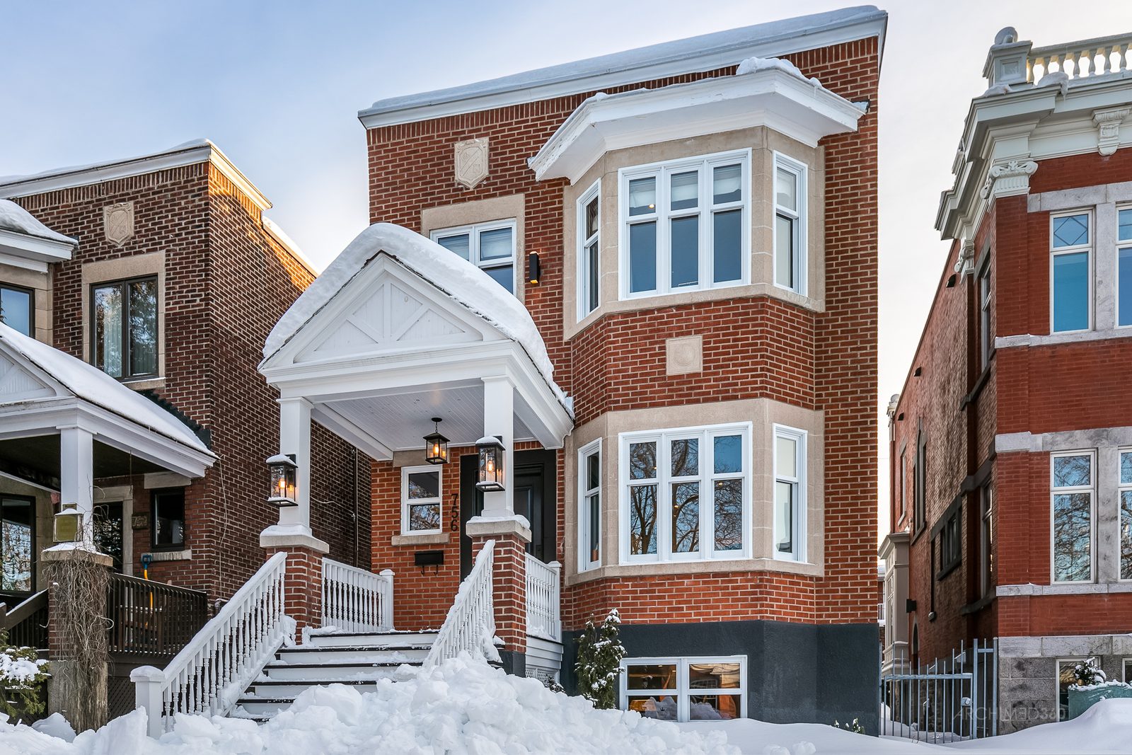 Maison de briques rouges avec fenêtres en baie et portique blanc, sur une rue résidentielle enneigée