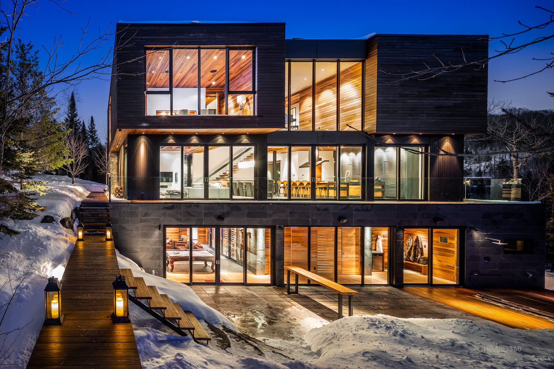 Façade vitrée illuminée un soir d'hiver, passerelle de bois et lanternes